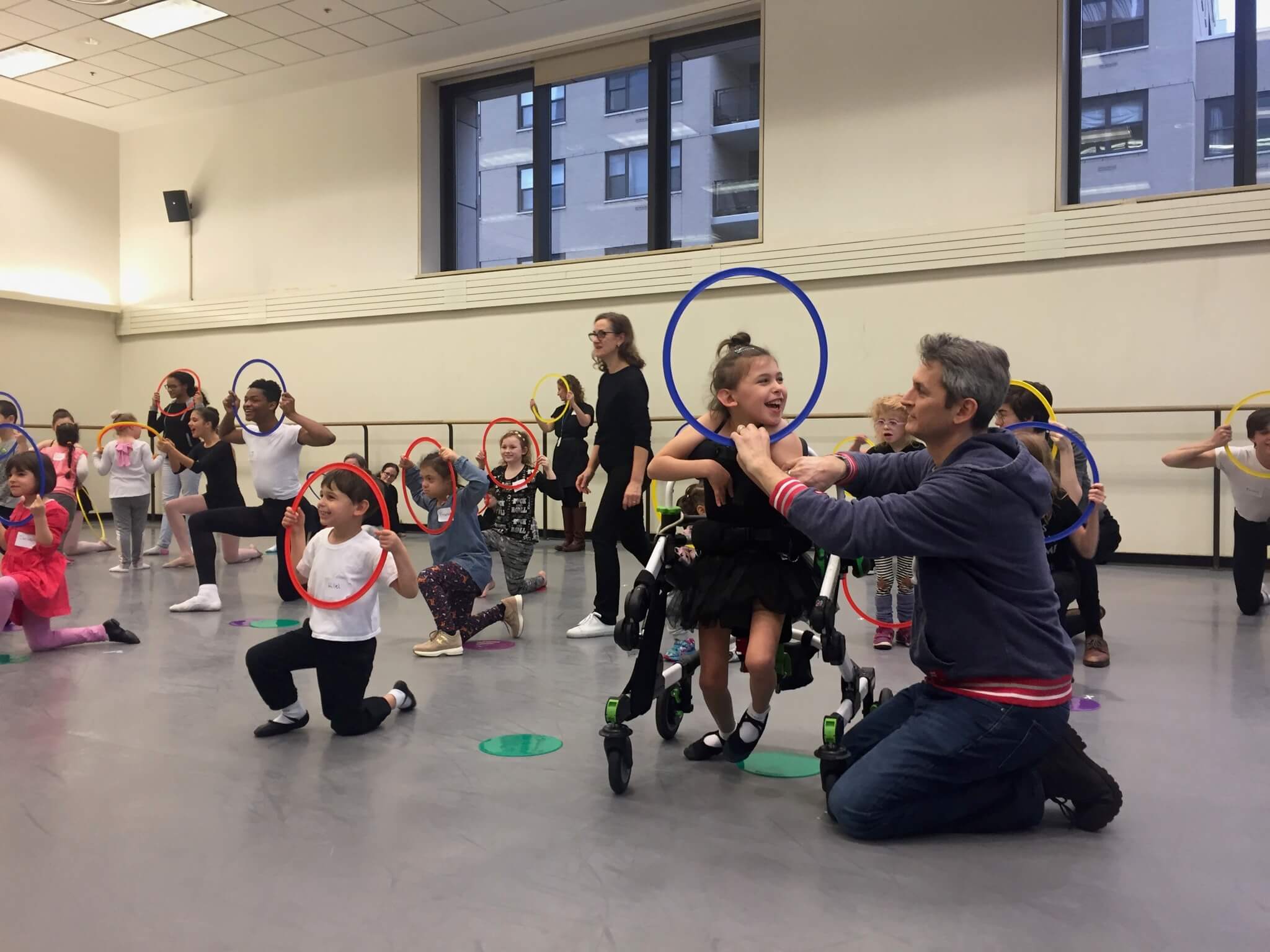Photo of a youth dance class, with the focus on one young girl who has a physical disability, smiling as an adult man assists her in participation by holding a ring to her face.