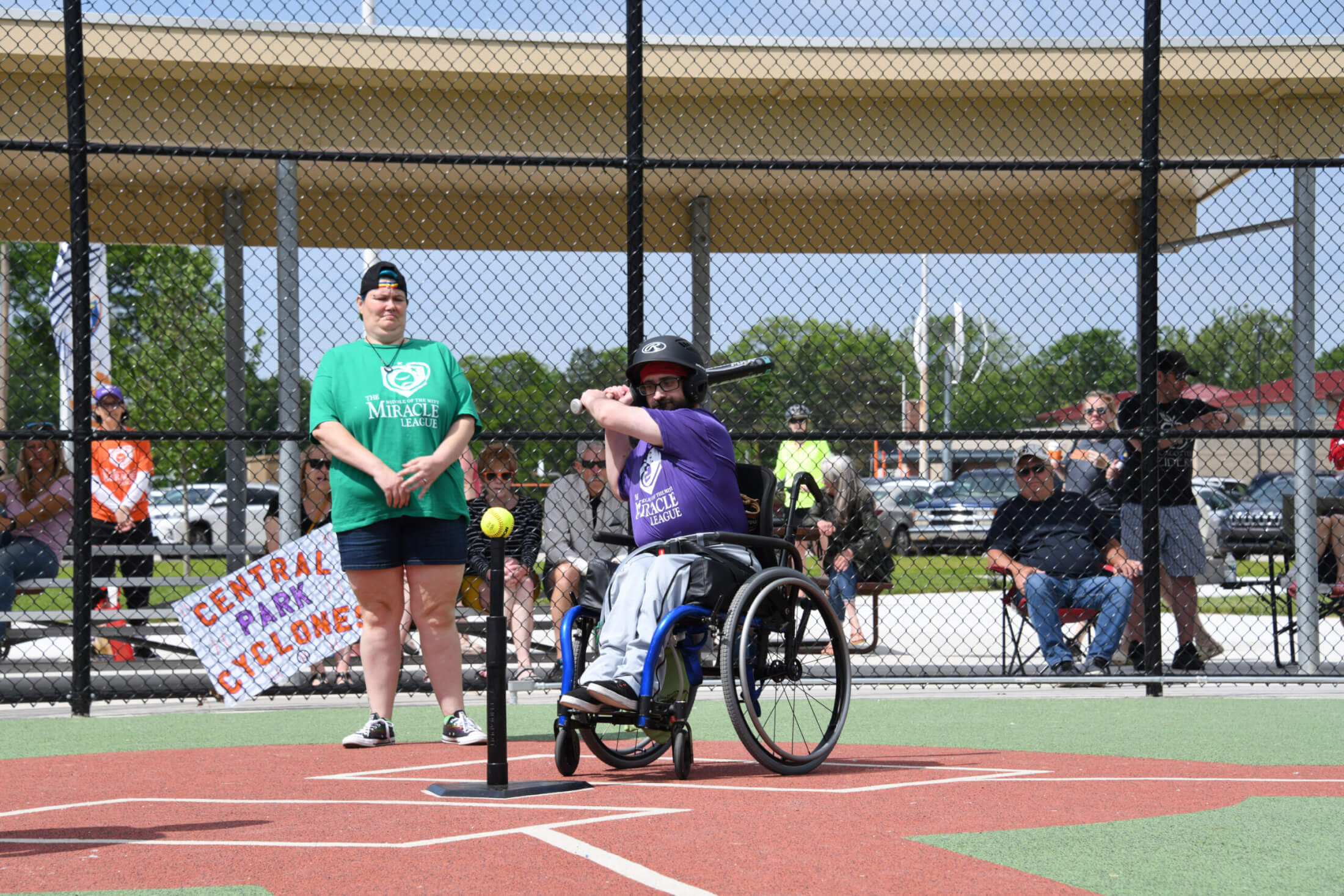 Photo of a man in a wheelchair hitting a baseball off a tee, as part of The Miracle League baseball program in Midland, Michigan.